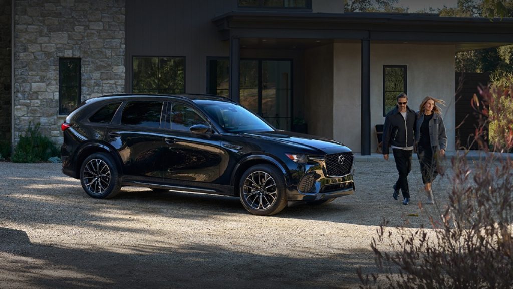A black Mazda CX-70 SUV parked in front of a modern home with two people walking towards it