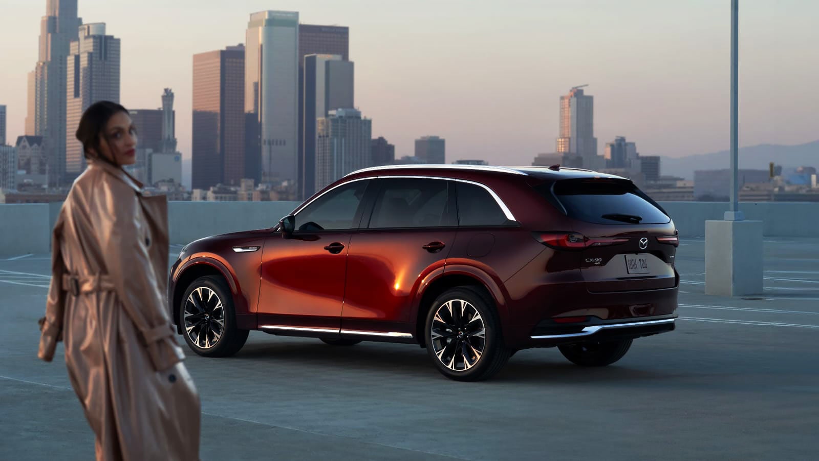 Red Mazda CX-90 SUV parked on a rooftop with a woman and city skyline at sunset.